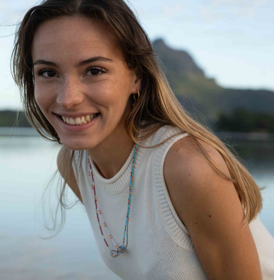 A smiling woman with long light brown hair wearing a white sleeveless knit top and a colorful beaded necklace, photographed outdoors against a soft-focus water and mountain background.