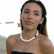 A woman wearing a chunky white beaded necklace with a small blue accent bead, photographed outdoors against a bright, overexposed background.