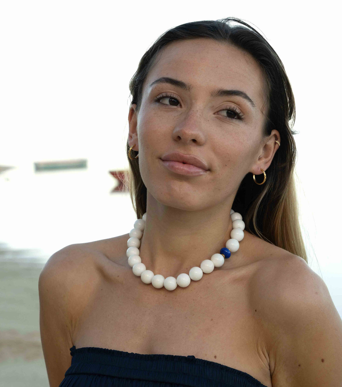 A woman wearing a chunky white beaded necklace with a small blue accent bead, photographed outdoors against a bright, overexposed background.
