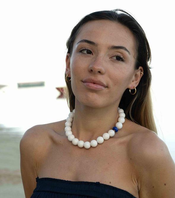A woman wearing a chunky white beaded necklace with a small blue accent bead, photographed outdoors against a bright, overexposed background.