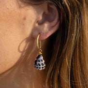 A close-up of a person's ear wearing a gold hoop earring with a teardrop-shaped shell charm featuring a black and white spotted pattern.