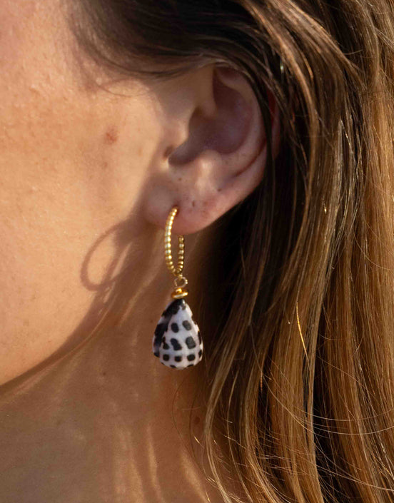 A close-up of a person's ear wearing a gold hoop earring with a teardrop-shaped shell charm featuring a black and white spotted pattern.