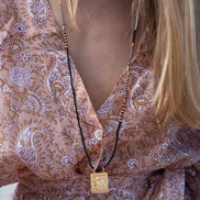 A close-up of a woman wearing a long black cord necklace with a square gold pendant, styled over a pink paisley patterned blouse.