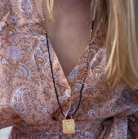 A close-up of a woman wearing a long black cord necklace with a square gold pendant, styled over a pink paisley patterned blouse.