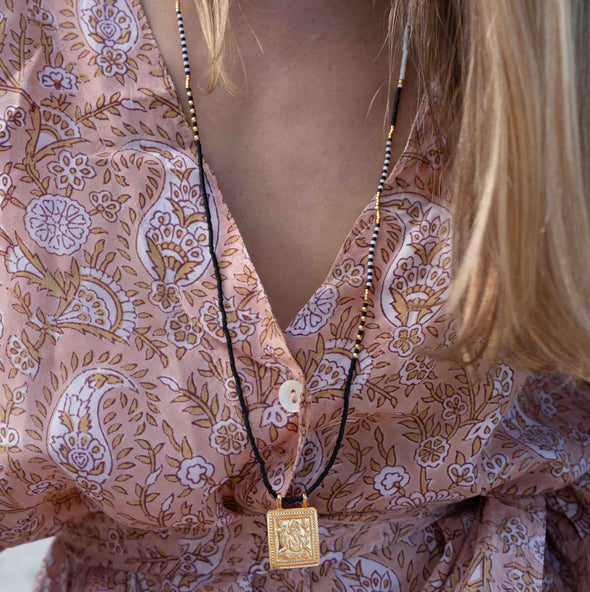 A close-up of a woman wearing a long black cord necklace with a square gold pendant, styled over a pink paisley patterned blouse.