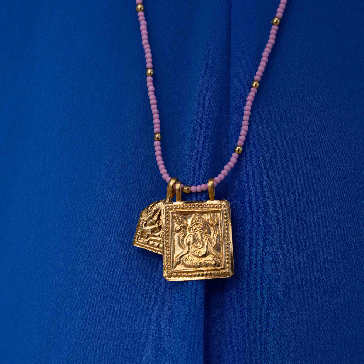 A gold-toned pendant necklace featuring an embossed Ganesha design, hanging from a strand of small pinkish-purple beads with tiny gold spacer beads, set against a vibrant blue fabric background.