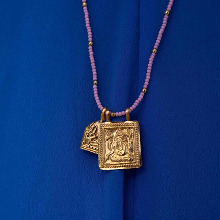 A gold-toned pendant necklace featuring an embossed Ganesha design, hanging from a strand of small pinkish-purple beads with tiny gold spacer beads, set against a vibrant blue fabric background.