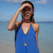 A woman standing on a beach wearing a bright blue halter dress and layered necklaces, with her hand raised to shield her eyes from the sun against a blue sky and ocean background.