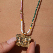 A close-up of a person's chest with a beaded necklace hanging from a black strap, held by a hand. The necklace features a gold-toned rectangular pendant, white beads, a pink strand, and a green woven section with small gold spacer beads.