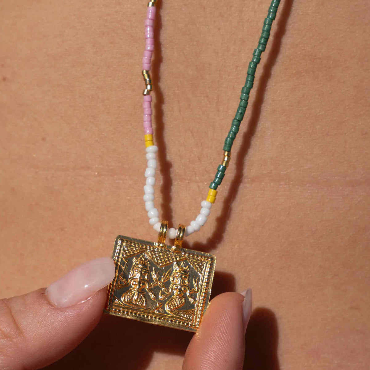 A close-up of a person's chest with a beaded necklace hanging from a black strap, held by a hand. The necklace features a gold-toned rectangular pendant, white beads, a pink strand, and a green woven section with small gold spacer beads.
