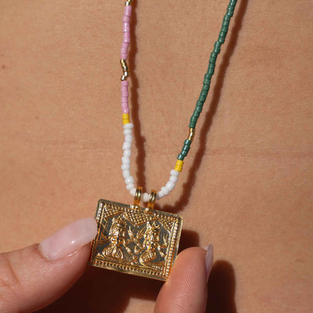 A close-up of a person's chest with a beaded necklace hanging from a black strap, held by a hand. The necklace features a gold-toned rectangular pendant, white beads, a pink strand, and a green woven section with small gold spacer beads.
