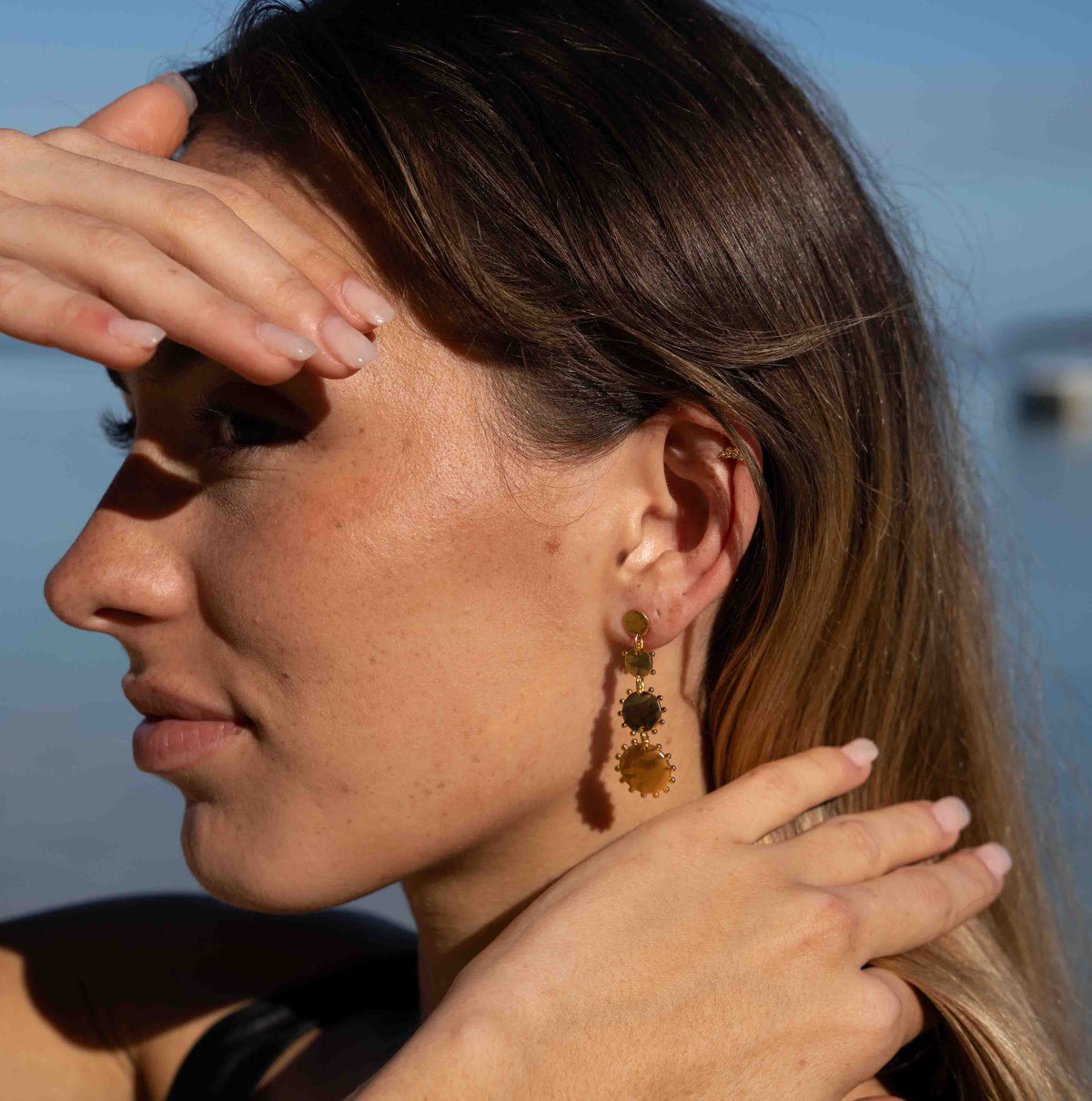 A woman in profile outdoors wearing a long, dangling statement earring made of stacked sunflower-like discs in olive green, dark green, and gold tones.