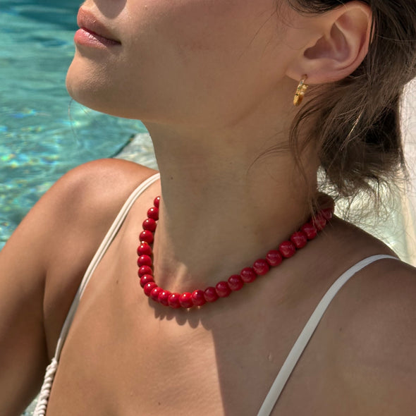 A close-up lifestyle shot of a woman wearing a chunky red beaded necklace and a small gold hoop earring, with a swimming pool in the background.