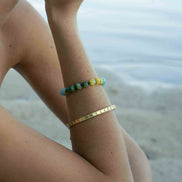 A person wearing two bracelets on their wrist at the beach: a gold-tone metal cuff bracelet with small engraved dots and a beaded bracelet with green, blue, and yellow beads.