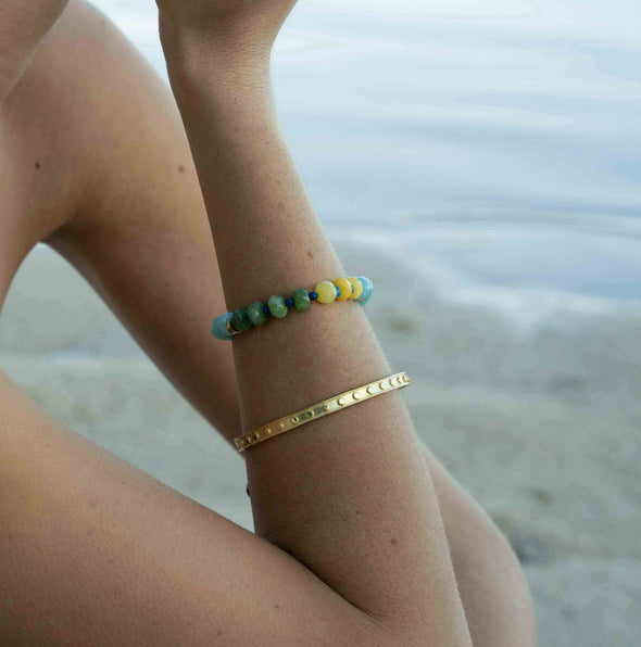 A person wearing two bracelets on their wrist at the beach: a gold-tone metal cuff bracelet with small engraved dots and a beaded bracelet with green, blue, and yellow beads.