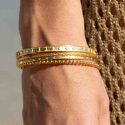 A close-up of a person's wrist wearing three stacked gold bangles, with one bangle featuring a row of small clear stones and another with a beaded border, against a neutral background.