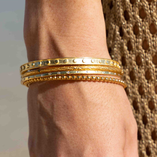 A close-up of a person's wrist wearing three stacked gold bangles, with one bangle featuring a row of small clear stones and another with a beaded border, against a neutral background.