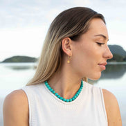 A woman in a white sleeveless top wearing a turquoise beaded necklace and a small gold hoop earring, photographed outdoors with a blurred lake and hills in the background.