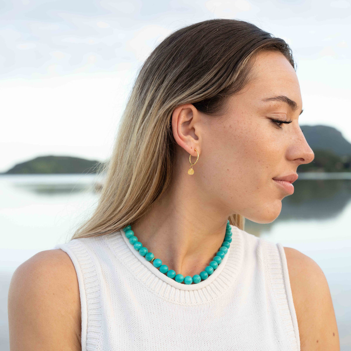 A woman in a white sleeveless top wearing a turquoise beaded necklace and a small gold hoop earring, photographed outdoors with a blurred lake and hills in the background.