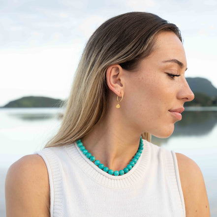 A woman in a white sleeveless top wearing a turquoise beaded necklace and a small gold hoop earring, photographed outdoors with a blurred lake and hills in the background.