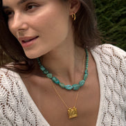 A close-up lifestyle shot of a woman wearing a turquoise beaded necklace and a gold square pendant necklace, with a white knit top and blurred green foliage in the background.