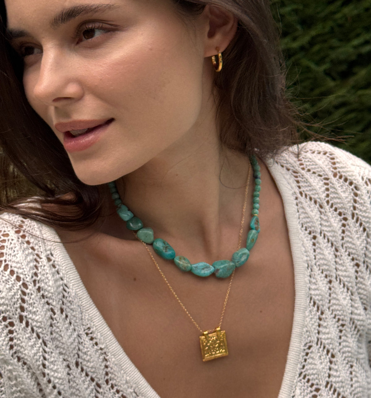 A close-up lifestyle shot of a woman wearing a turquoise beaded necklace and a gold square pendant necklace, with a white knit top and blurred green foliage in the background.