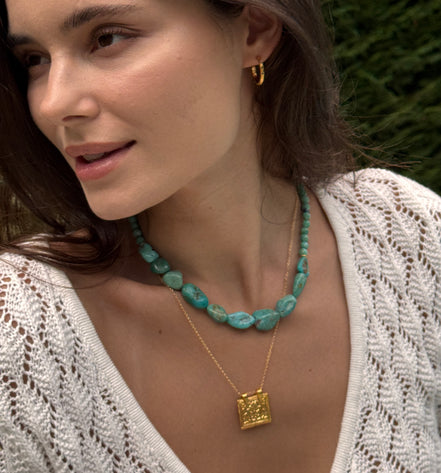 A close-up lifestyle shot of a woman wearing a turquoise beaded necklace and a gold square pendant necklace, with a white knit top and blurred green foliage in the background.