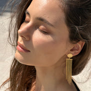A close-up of a woman wearing a gold dangle earring with a circular stud and multiple hanging chain strands, photographed outdoors with bright sunlight and a blurred beach-like background.