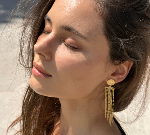 A close-up of a woman wearing a gold dangle earring with a circular stud and multiple hanging chain strands, photographed outdoors with bright sunlight and a blurred beach-like background.