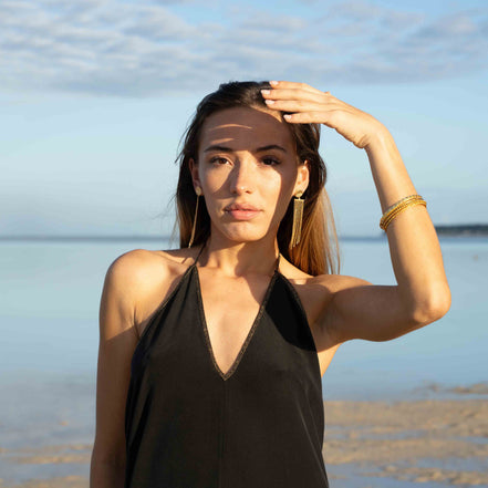 A woman posing on a beach wearing a black halter-neck top and gold jewelry.