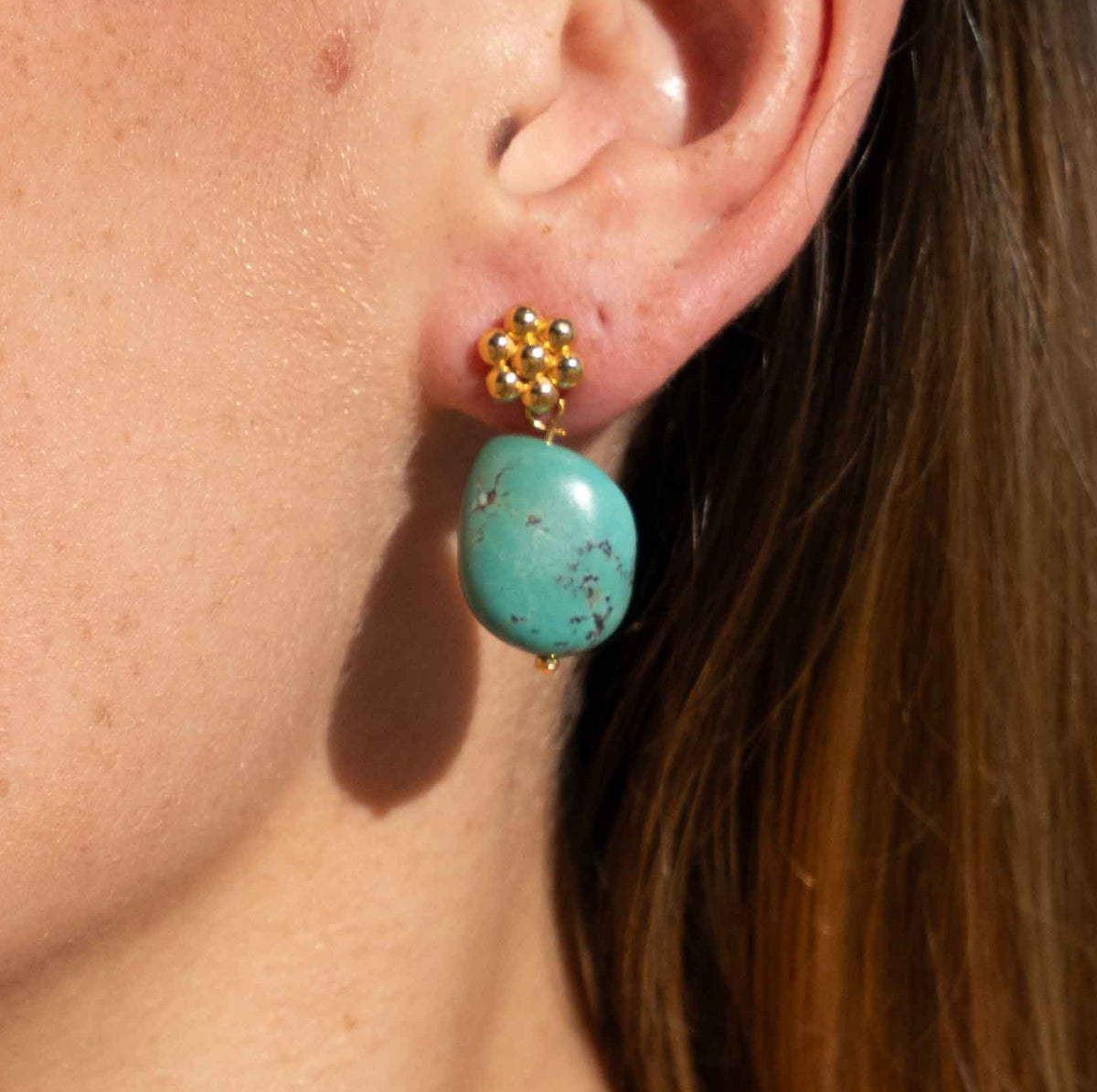 A close-up of a person's ear wearing a gold stud earring with a round turquoise drop pendant.