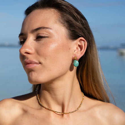 A woman with long brown hair is shown in profile outdoors near water, wearing a delicate gold chain necklace and a matching gold drop earring with a round turquoise-blue stone.